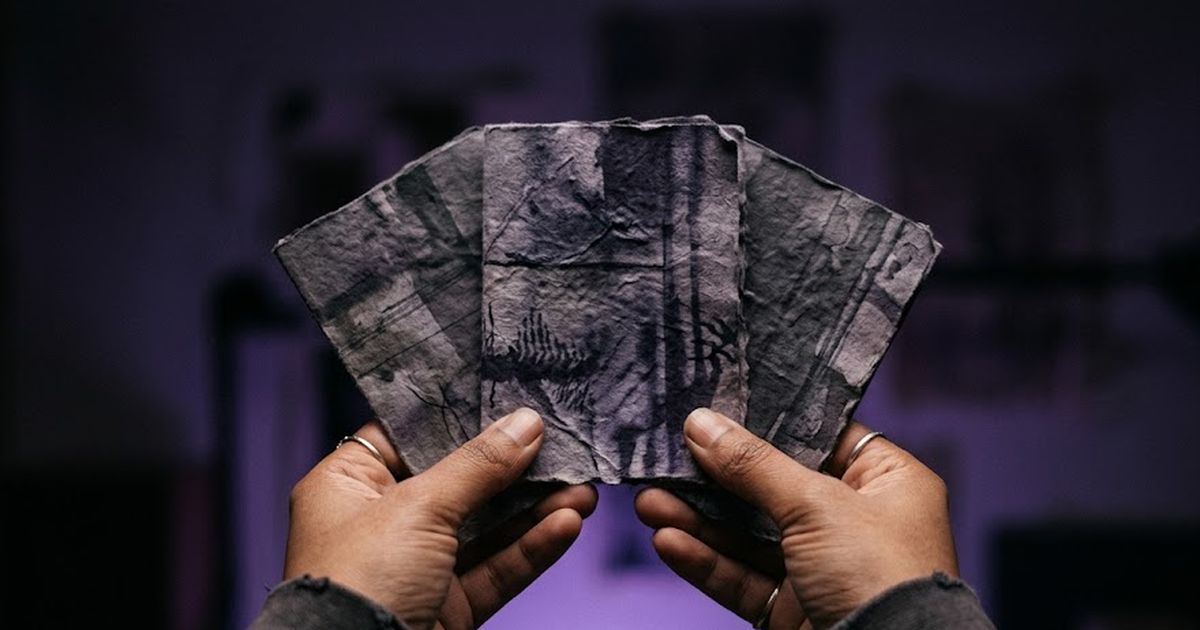 South Asian hands holding three small cards fanned out, choosing one, dark purple background